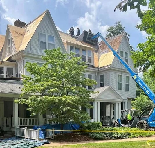 Blue crane lifts workers repairing roof of white three-story house